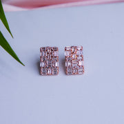 A pair of rose gold rectangle-shaped everyday silver stud earrings with sparkling embellishments, placed on a blue surface next to a green leaf.